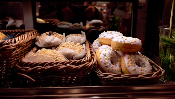 A display of pastries in wicker baskets, featuring a variety of crumb-topped muffins on the left and white frosted donuts with colorful round sprinkles on the right.